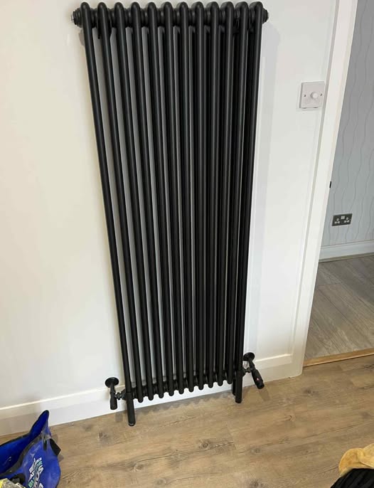 Tall black column radiator fitted to a white wall with valves and pipe connections at floor level