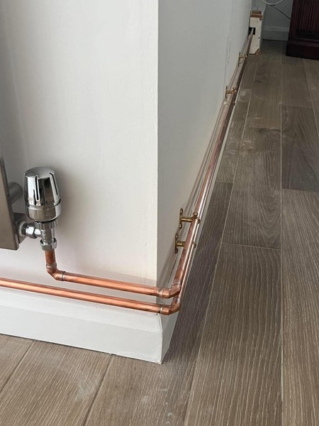Copper heating pipes following a skirting board around a wall corner, connected to a chrome radiator valve