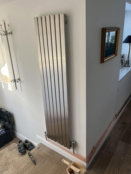 Tall silver-coloured flat-panel radiator on a white wall with chrome valves and copper pipework at skirting-board level